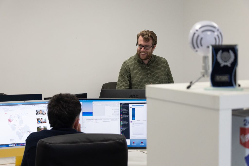 An Intimation designer smiling across the desks in the Newcastle studio, with an industry award on the shelf beside him