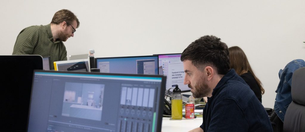 The Intimation studio in Newcastle: a designer cutting video on a large monitor with the timeline open, and colleagues working at desks behind