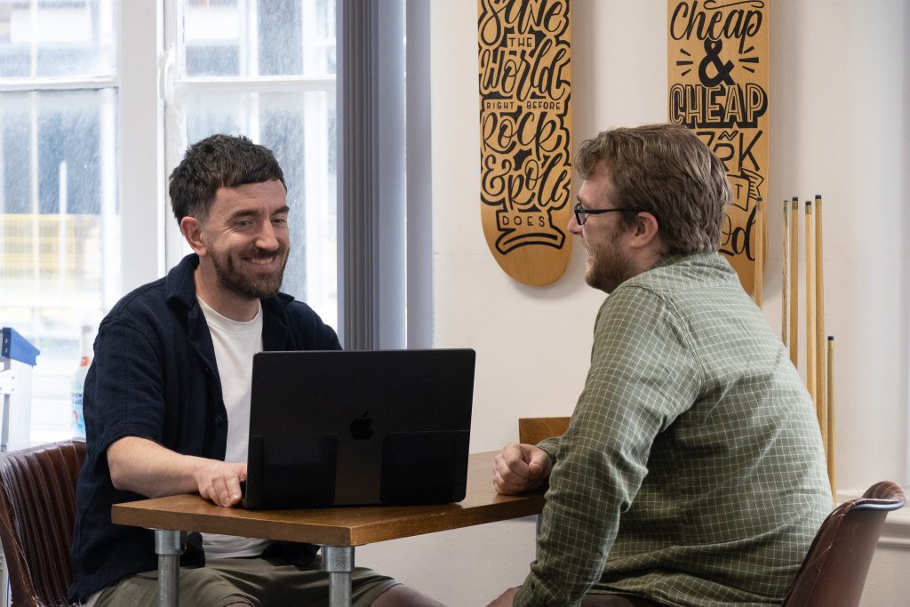 Two Intimation colleagues sitting at a table in the Newcastle studio, talking and smiling over an open laptop, with hand-lettered skateboard decks and pool cues on the wall behind them